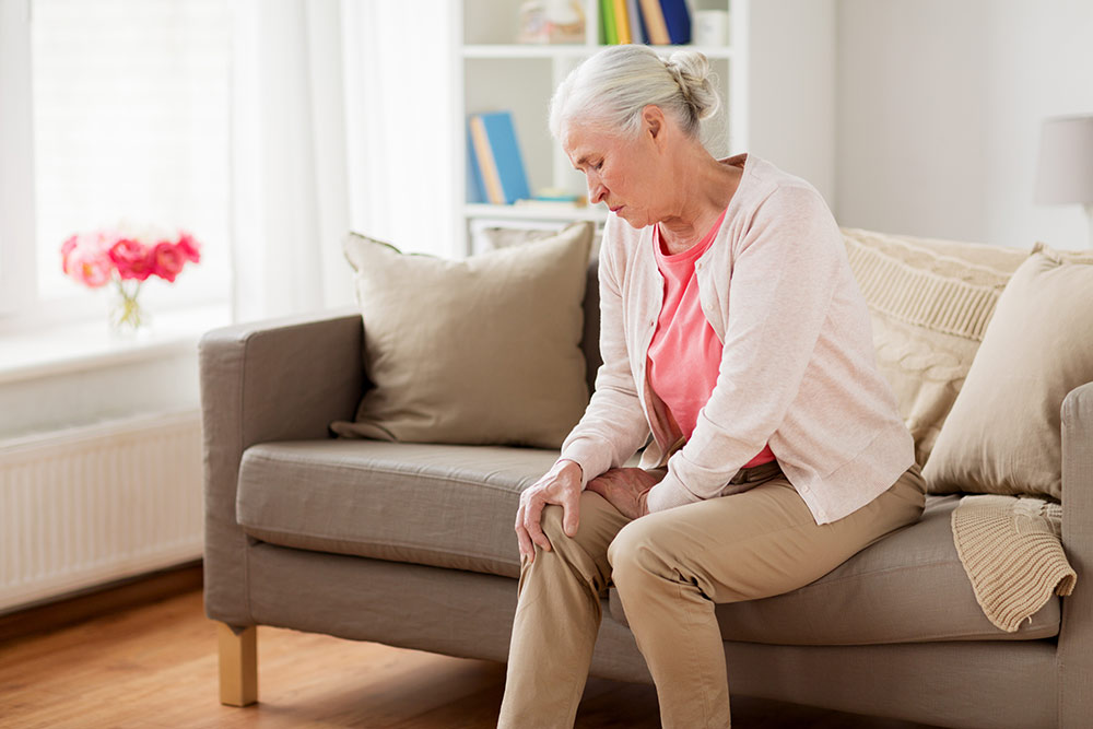 Senior woman sitting on couch holding knee, knee pain, arthritis