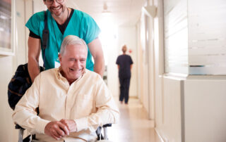 A senior man in a wheelchair going down the hallway with the staff at his skilled nursing facility